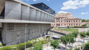 Un edificio moderno con fachada de vidrio y un patio con árboles y un edificio de ladrillo al fondo.