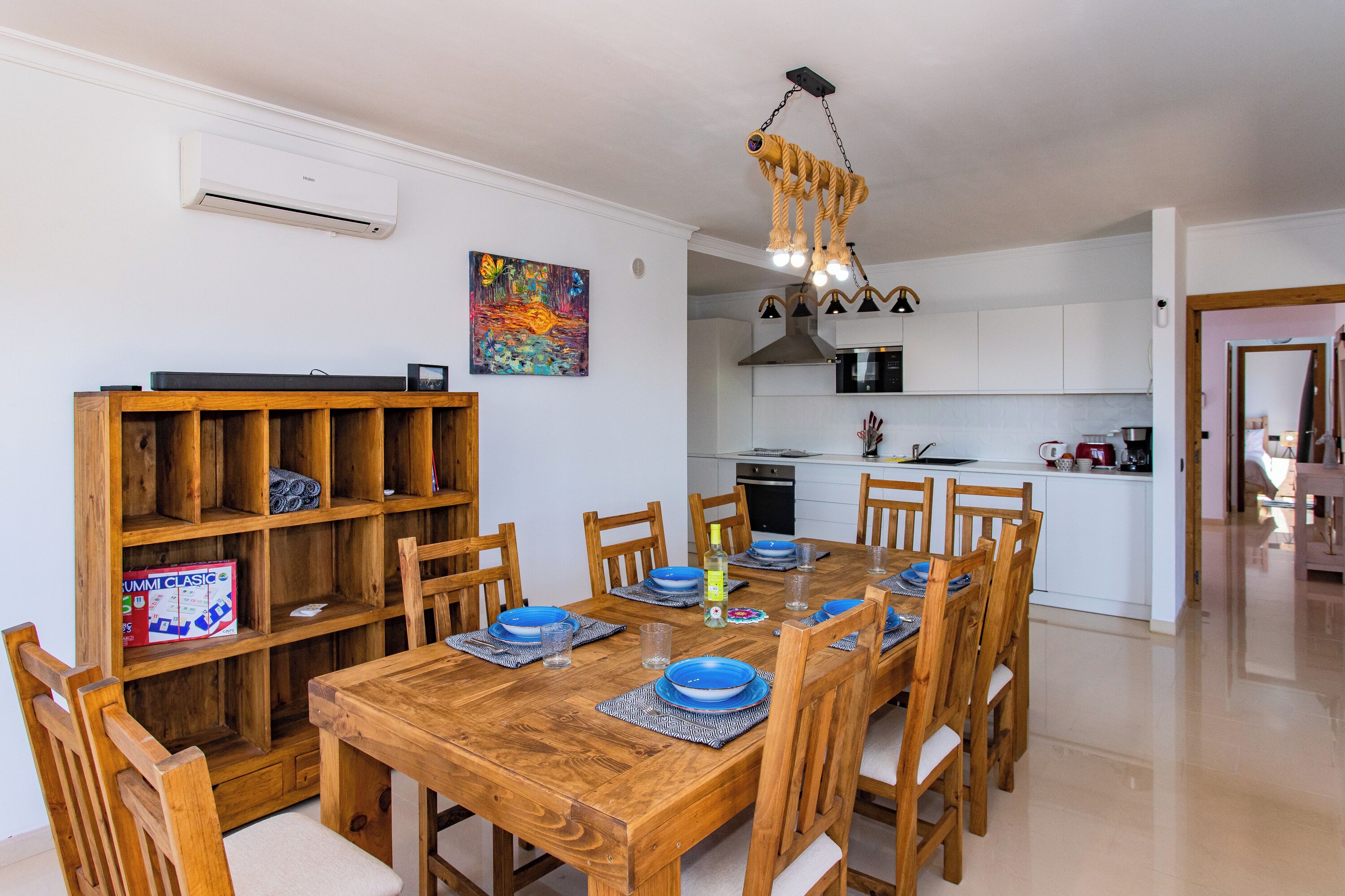 Een eetruimte met een houten tafel gedekt voor zes personen, omringd door houten stoelen. Links bevindt zich een boekenkast, en er is een keuken zichtbaar op de achtergrond.