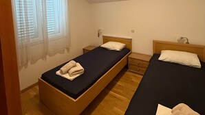 Una habitación con dos camas individuales, piso de madera, una ventana con cortinas ligeras y una mesita de noche con lámpara.