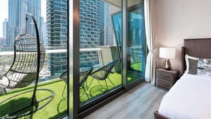 Et moderne hotelværelse med en stor skydedør, der fører ud til en balkon med udsigt til høje bygninger. Der er en seng med sengegavl, et natbord med lampe, og to stole på balkonen.