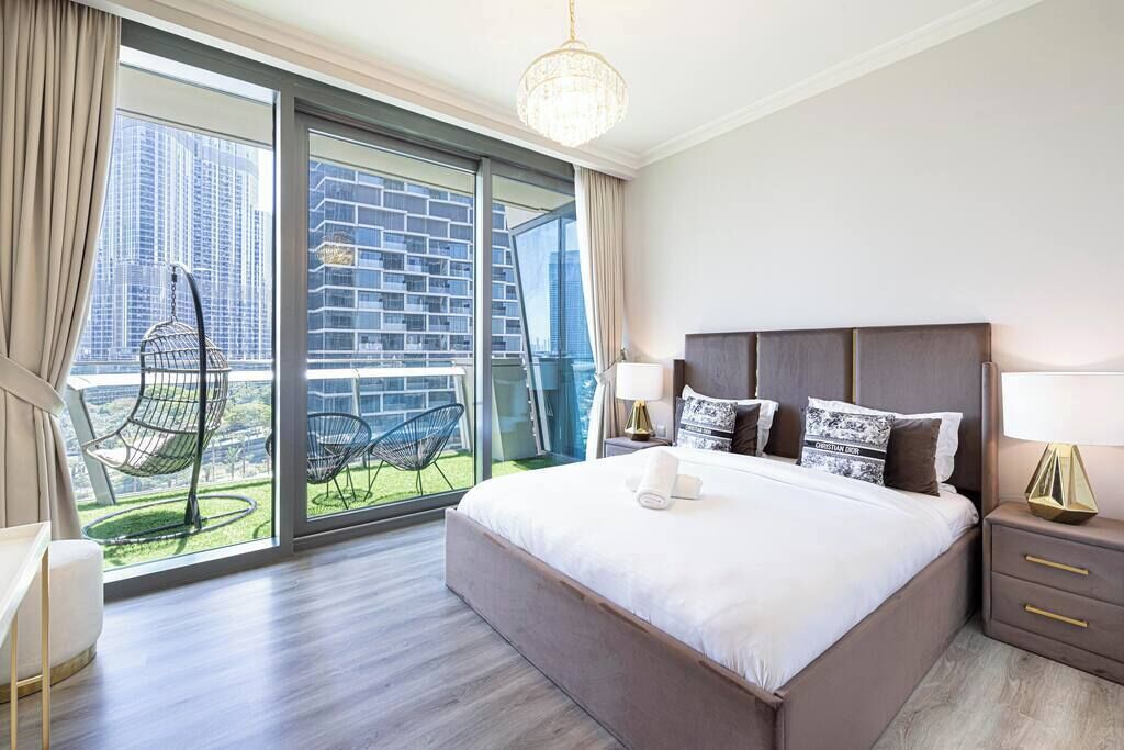Quarto de hotel moderno com uma cama grande, mesa de cabeceira, abajur e vista da cidade através de janelas do chão ao teto.