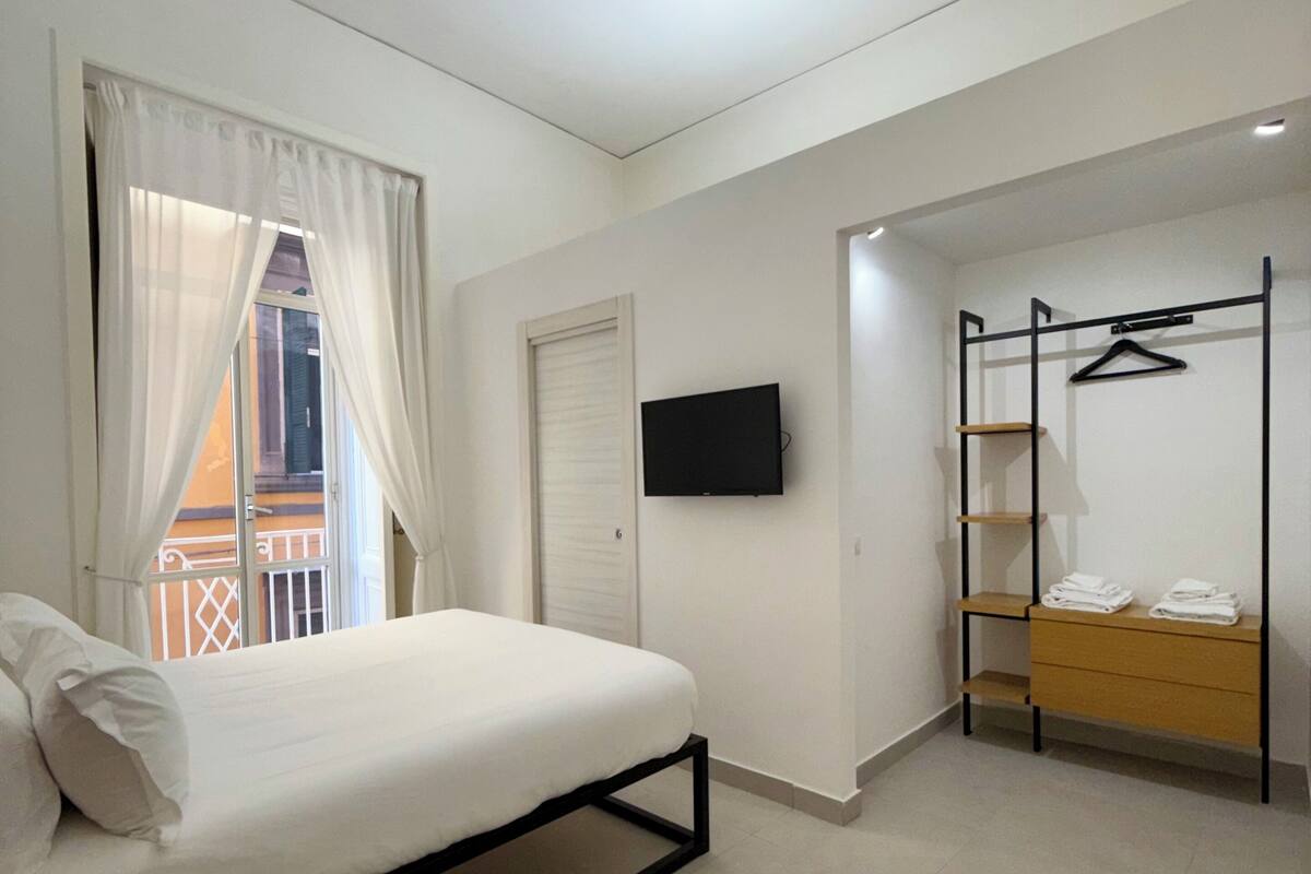 A bedroom with a bed, a TV, a closet with a clothing rack, and a balcony view.