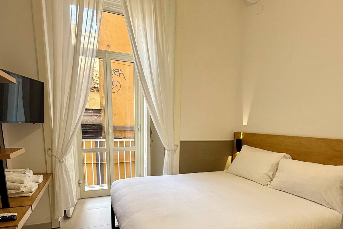 A hotel room with a bed, a TV, a bedside table, a window with curtains, and an air conditioning unit.