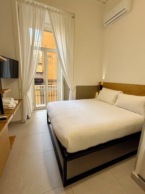 A hotel room with a bed, a TV, a bedside table, a window with curtains, and an air conditioning unit. - Nabab-Napoli Bed and Breakfast (Naples)