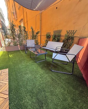 An outdoor seating area with artificial grass, metal chairs, and a table, surrounded by potted plants and a yellow wall. - Nabab-Napoli Bed and Breakfast (Naples)