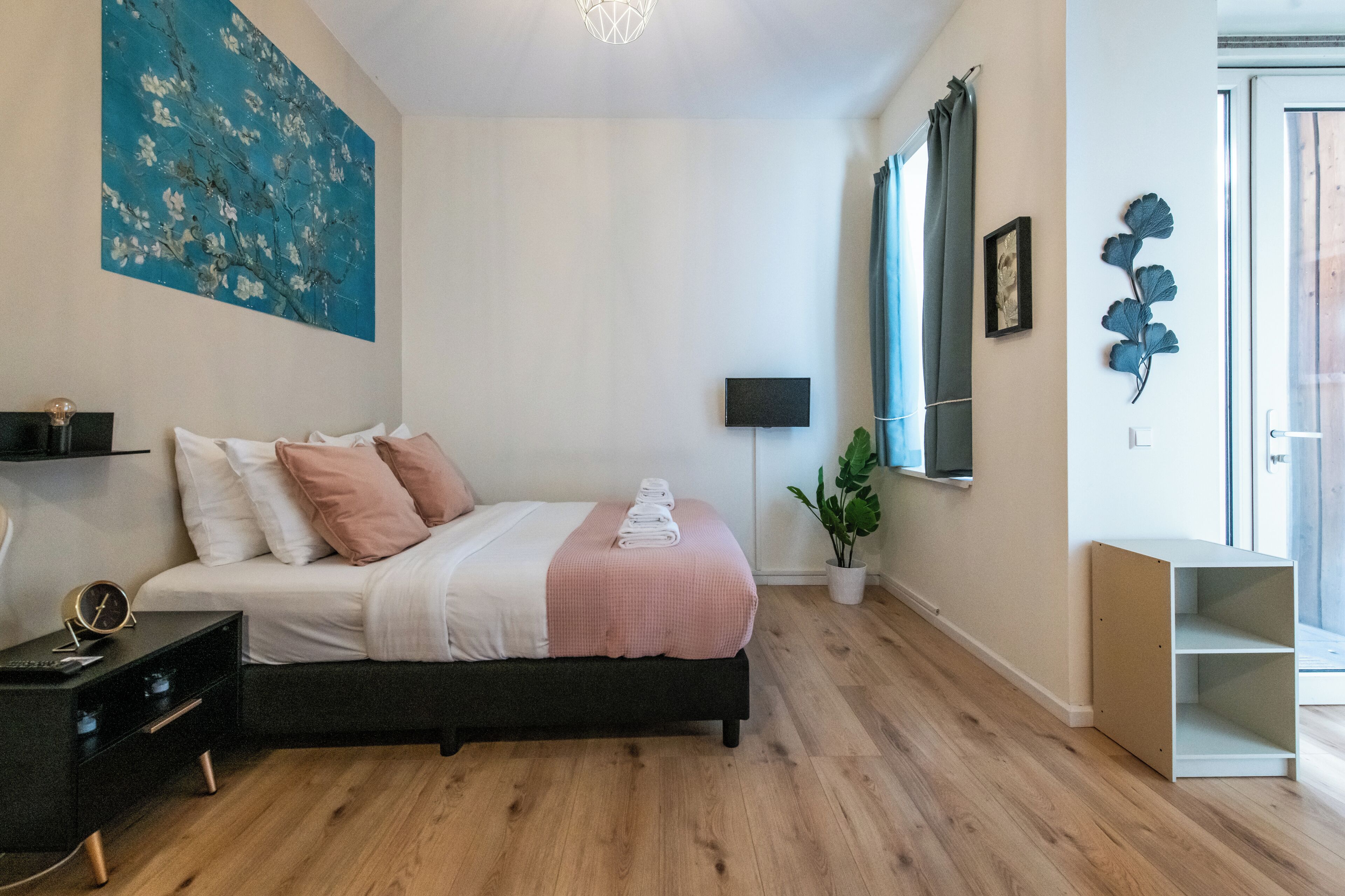 A bedroom with a bed, bedside table, lamp, artwork, and a window with curtains.