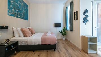 Une chambre à coucher comprenant un lit, une table de chevet, une lampe, un tableau et une fenêtre avec des rideaux.