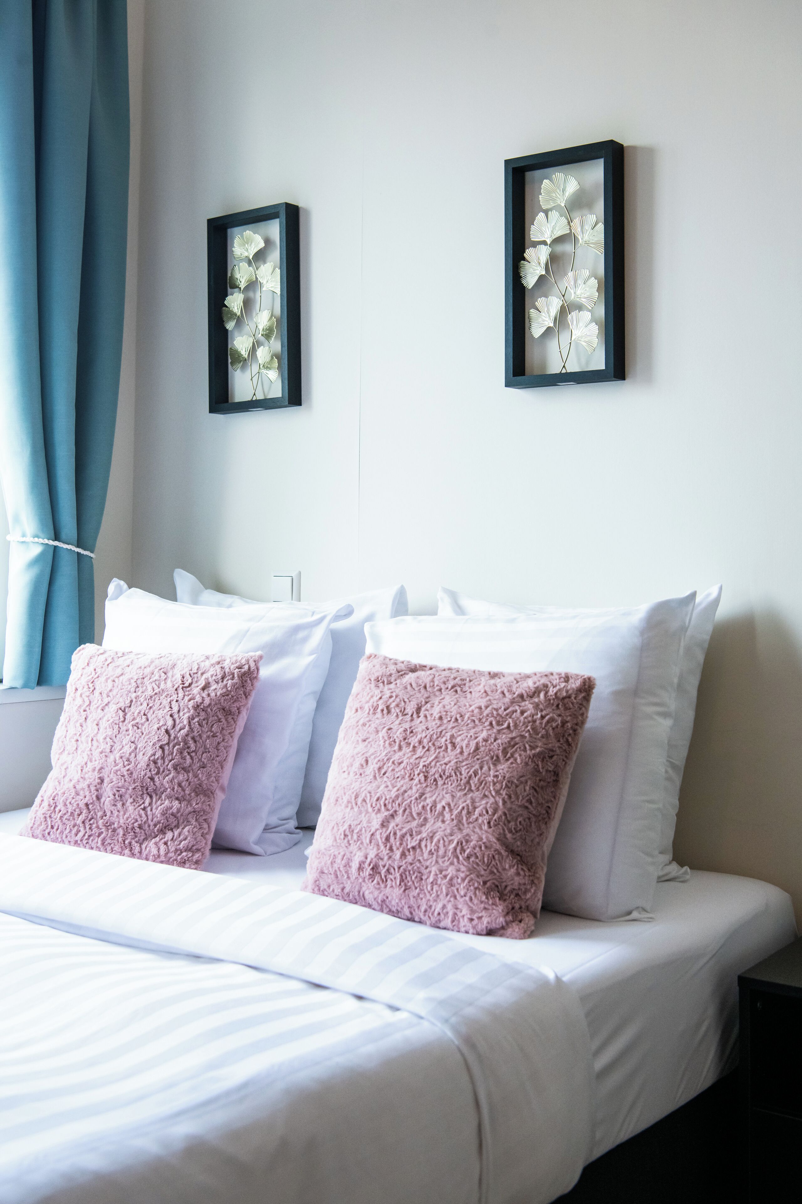 Una cama bien hecha con almohadas blancas y rosas, una obra de arte botánica enmarcada en la pared, y una ventana con cortinas azules.