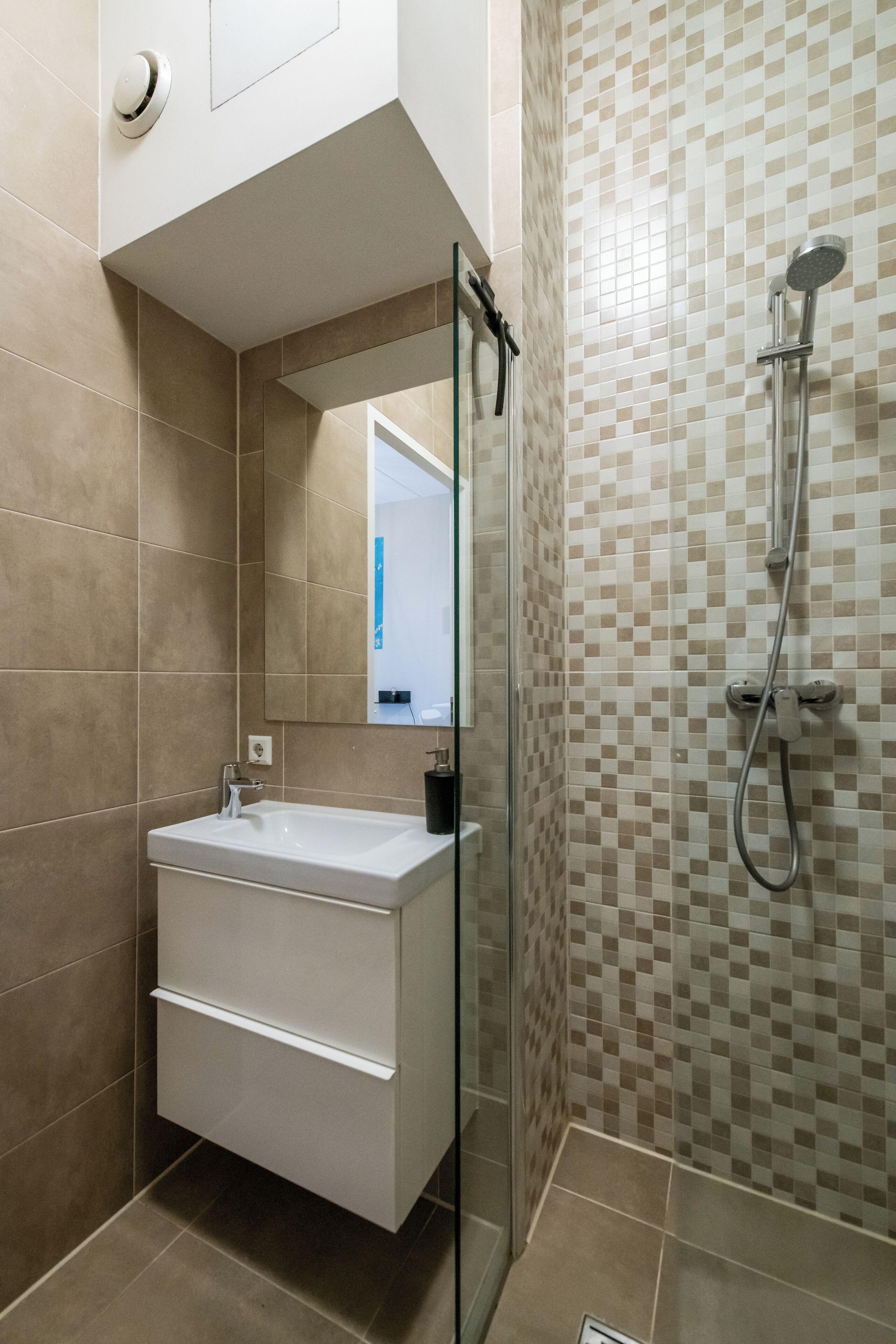 A modern bathroom with a glass-enclosed shower, a white sink with a mirror, and beige checkered tiles.