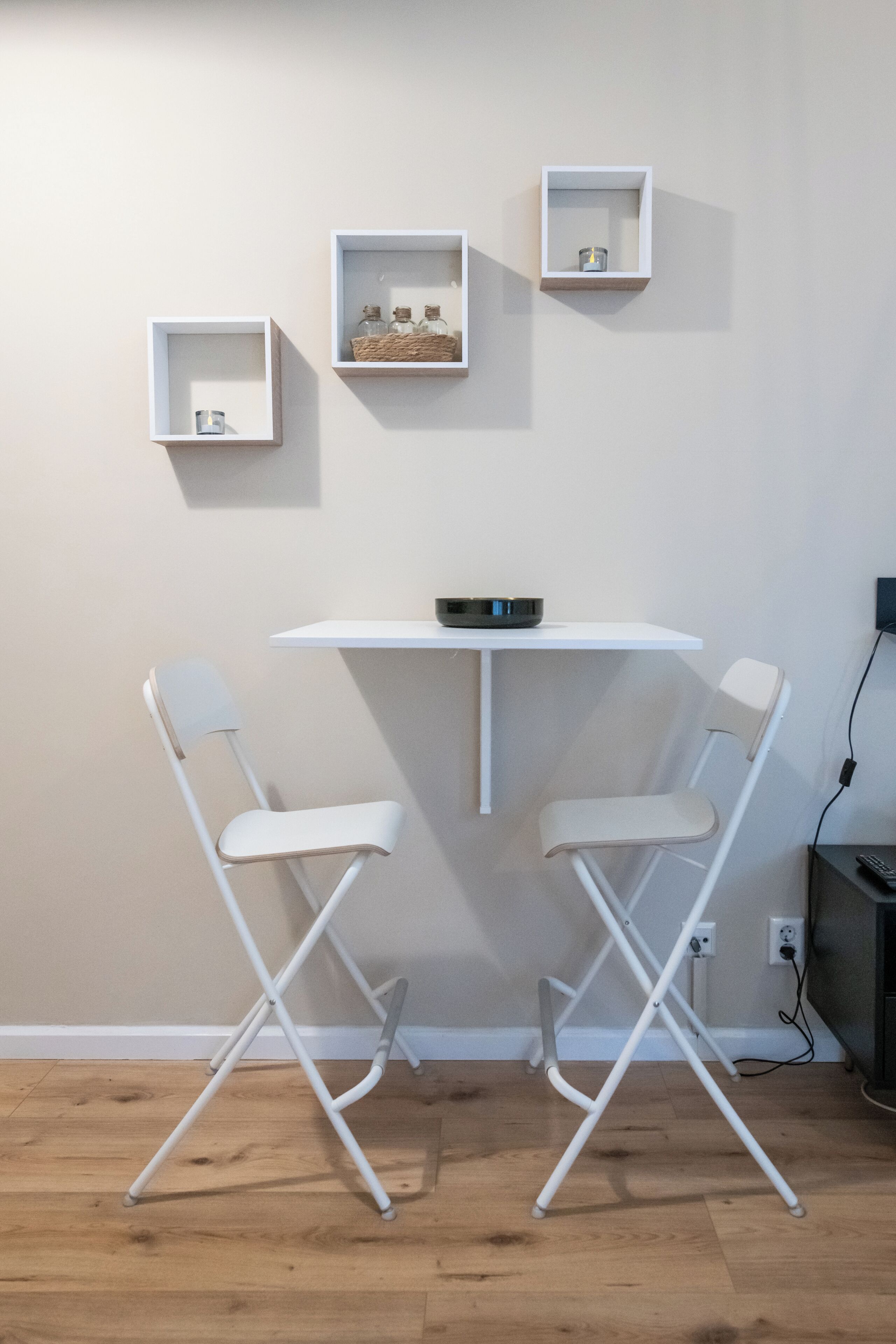 A small room with a white table and two white chairs. There are three white shelves on the wall, each containing different items.