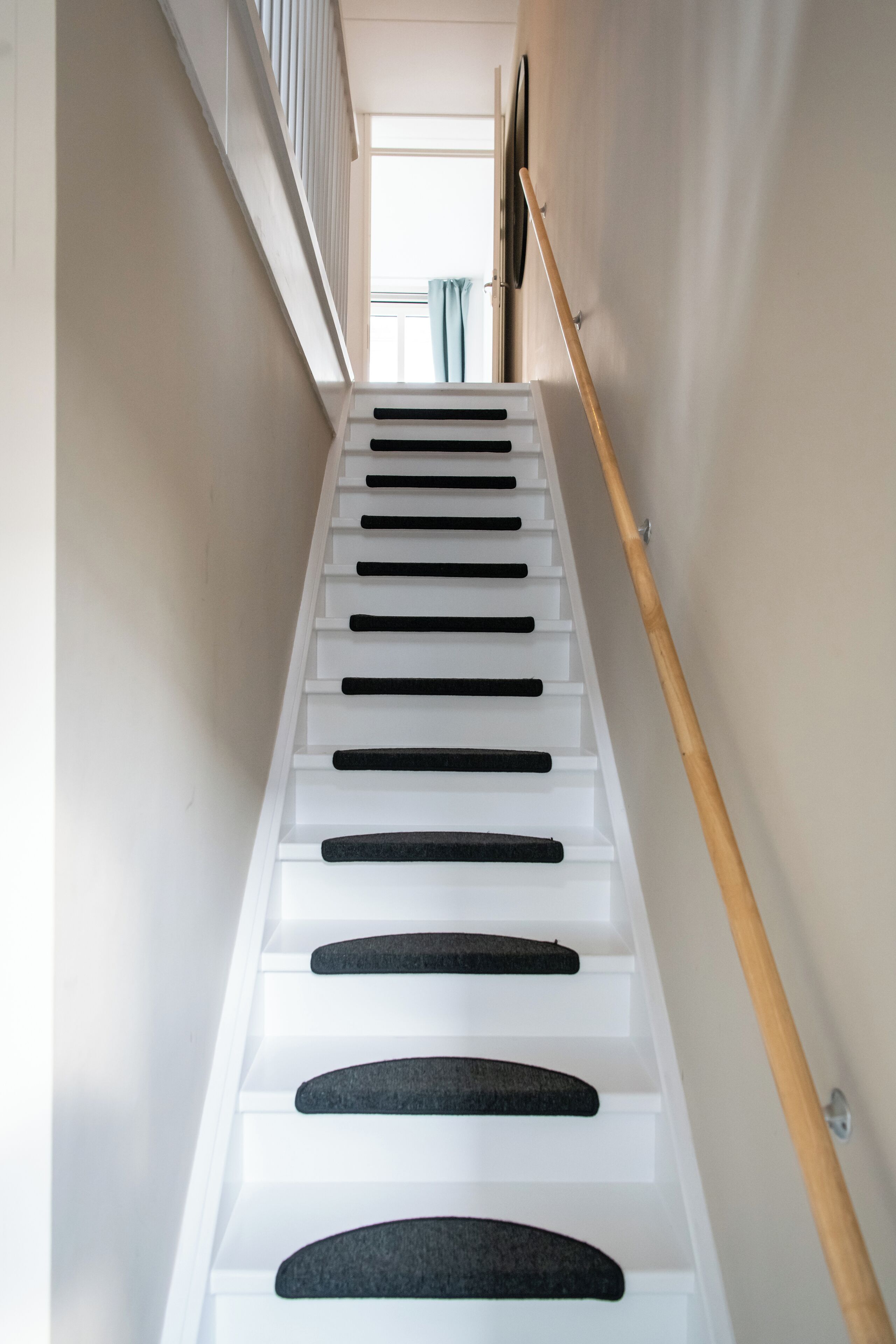 Un escalier avec des marches blanches et des bandes antidérapantes noires, menant à une pièce dotée d’une fenêtre et de rideaux.