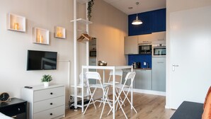 Un espacio habitable moderno y compacto con kitchenette, un comedor con mesa y sillas blancas, un televisor montado en la pared y una cama con mesita de noche.