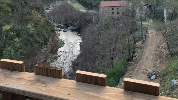 En träveranda med utsikt över en flod, omgiven av träd och en stenbro.