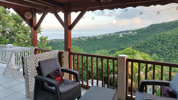 Zona relax all'aperto coperta, con sedie in vimini e un tavolo, affacciata su un paesaggio verdeggiante e una vista lontana del mare.