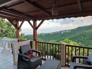 A covered outdoor seating area with wicker chairs and a table, overlooking a lush green landscape and a distant view of the sea.