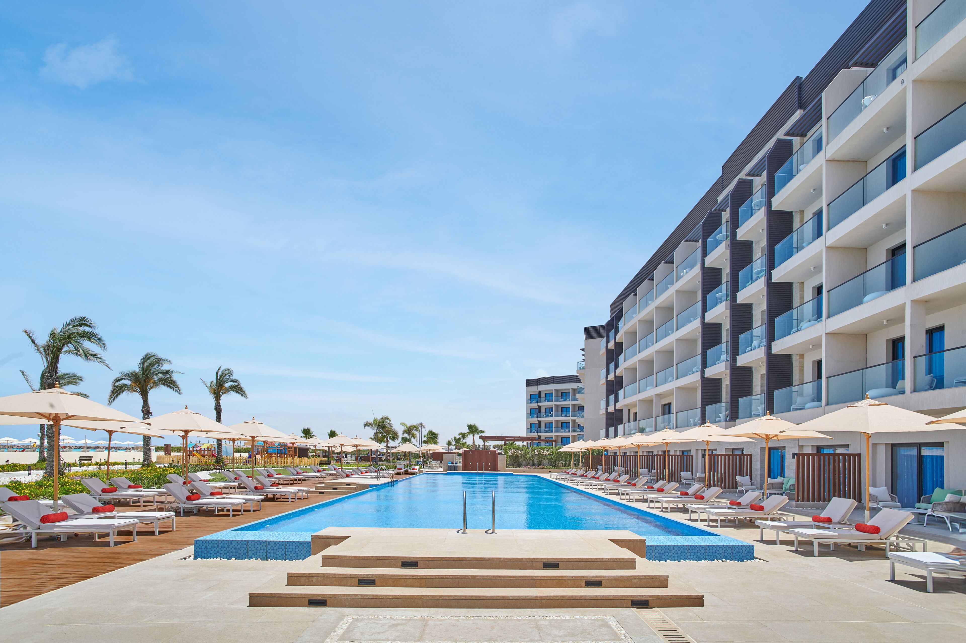 A hotel pool area with sun loungers and umbrellas, a clear sky, and a modern building with balconies.
