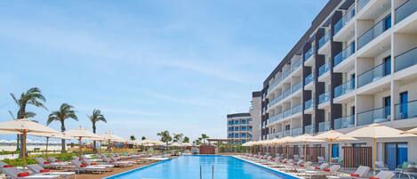 Área de piscina de hotel com espreguiçadeiras e guarda-sóis, céu claro e um edifício moderno com varandas.