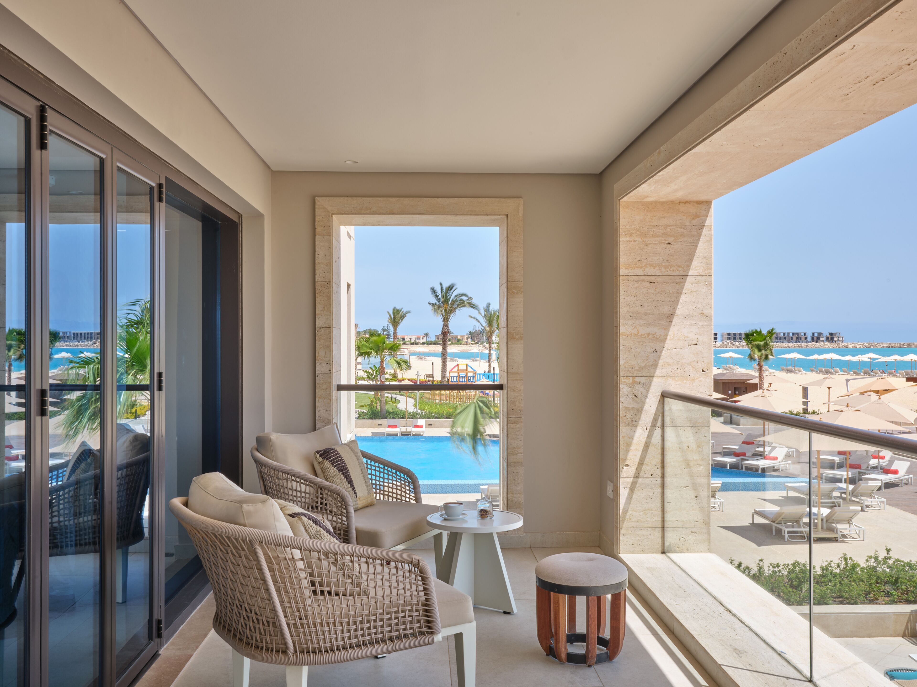 Un balcón con mobiliario de mimbre, una pequeña mesa redonda y vistas a una piscina y una playa.