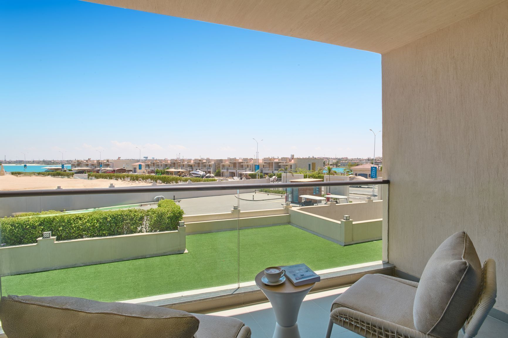 Un balcón con vista a la playa y al paisaje urbano, una mesa con un libro y una taza, y una silla de mimbre.