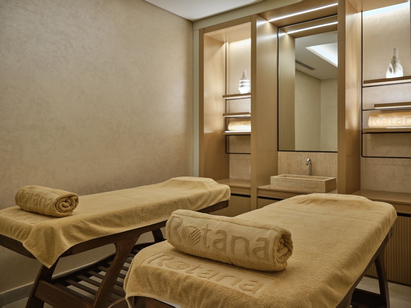Un salón de spa con dos camillas de masaje, cada una cubierta con una toalla color crema, y un espejo grande que refleja el interior de la habitación.
