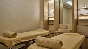 Un salón de spa con dos camillas de masaje, cada una cubierta con una toalla color crema, y un espejo grande que refleja el interior de la habitación.