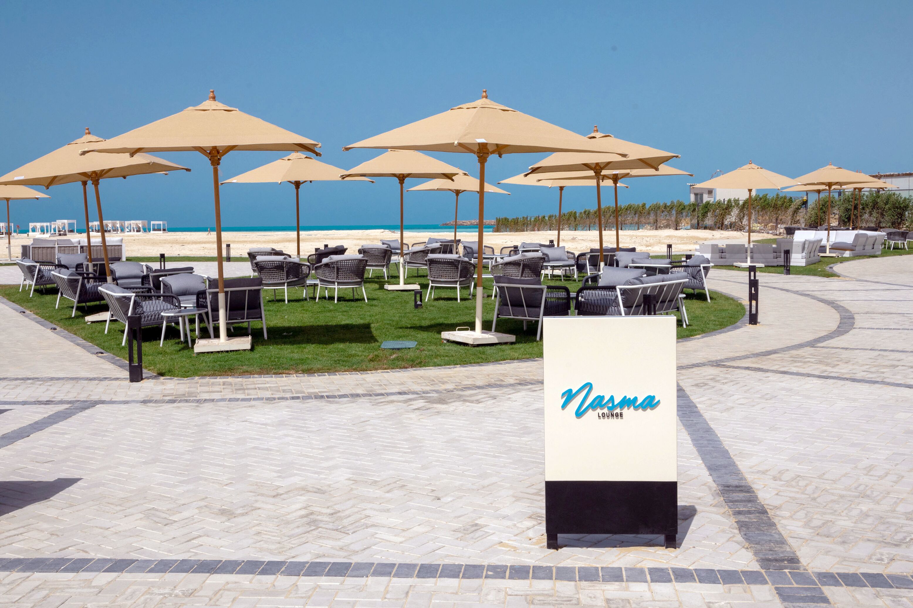 A beachside lounge area with multiple lounge chairs and umbrellas, a signboard with the name 'Nasma Lounge', and a clear view of the ocean.