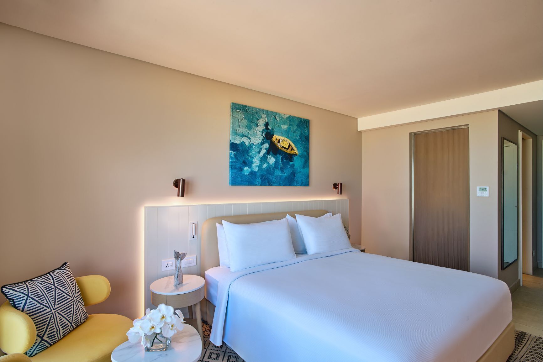A hotel room with a large bed, a small round table with a vase of flowers, a chair, and a painting on the wall.