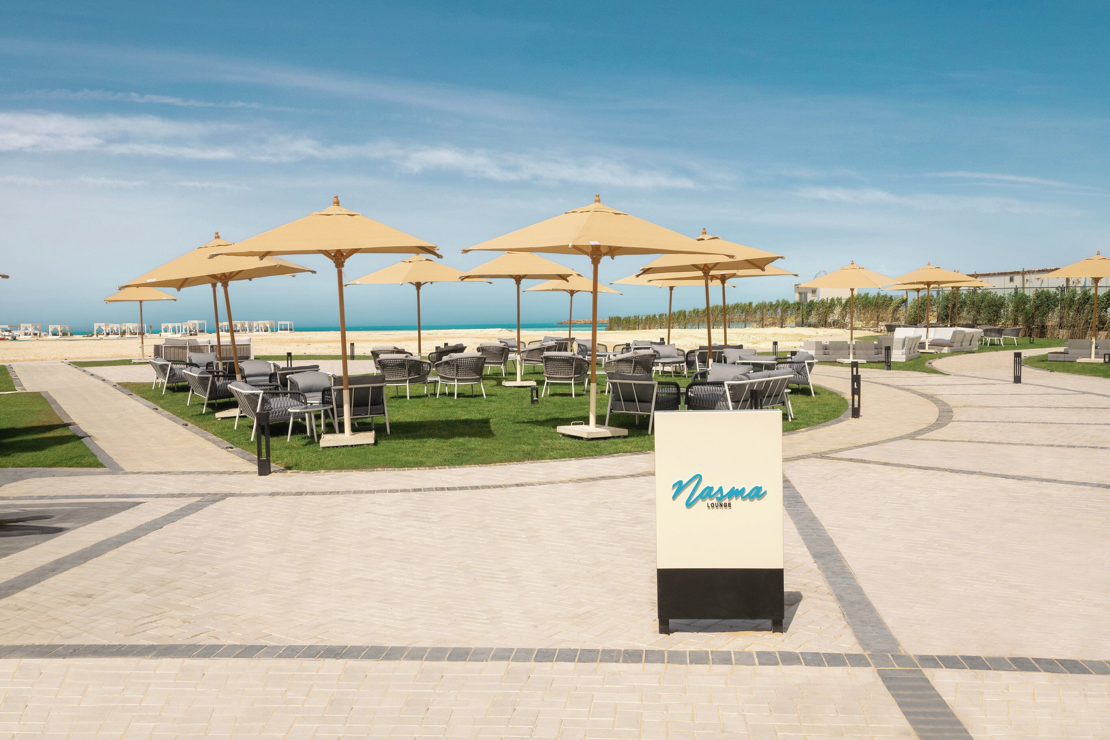 A beachside lounge area with multiple seating arrangements under large umbrellas, a sign reading "Nasmina LOUNGE," and a clear view of the ocean.