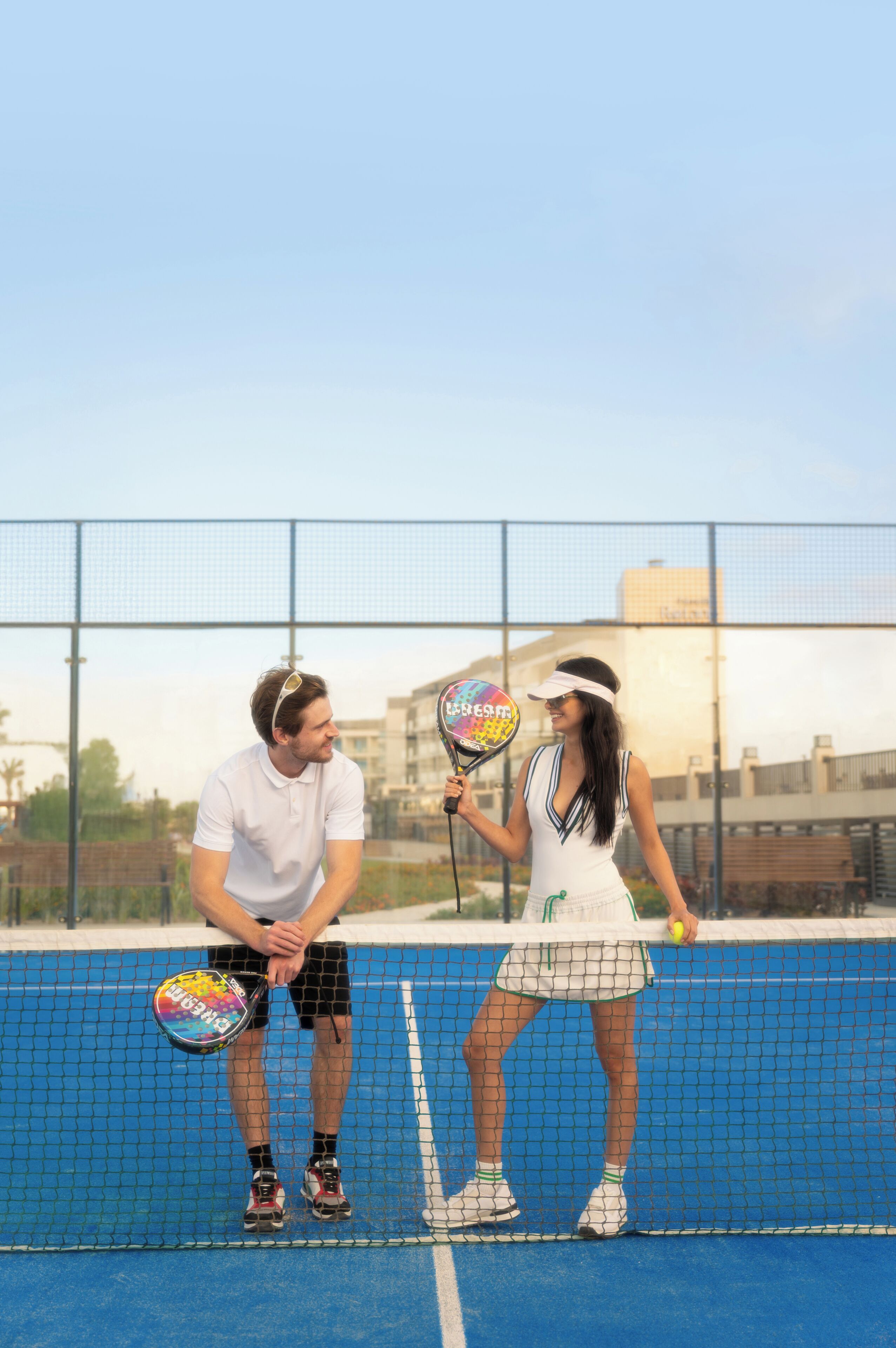 Deux personnes sur un court de tennis, avec un filet au milieu, une clôture en arrière-plan et des bâtiments au loin.
