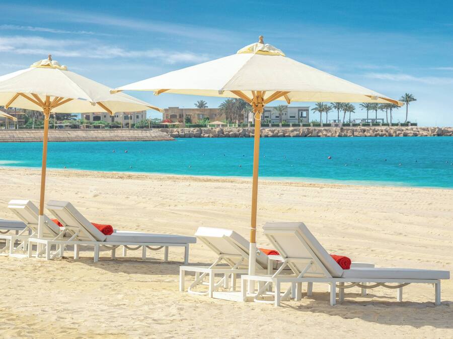 Une plage avec des chaises longues blanches et des parasols, une mer d’un bleu limpide et une côte rocheuse, avec des bâtiments visibles à l’arrière-plan.