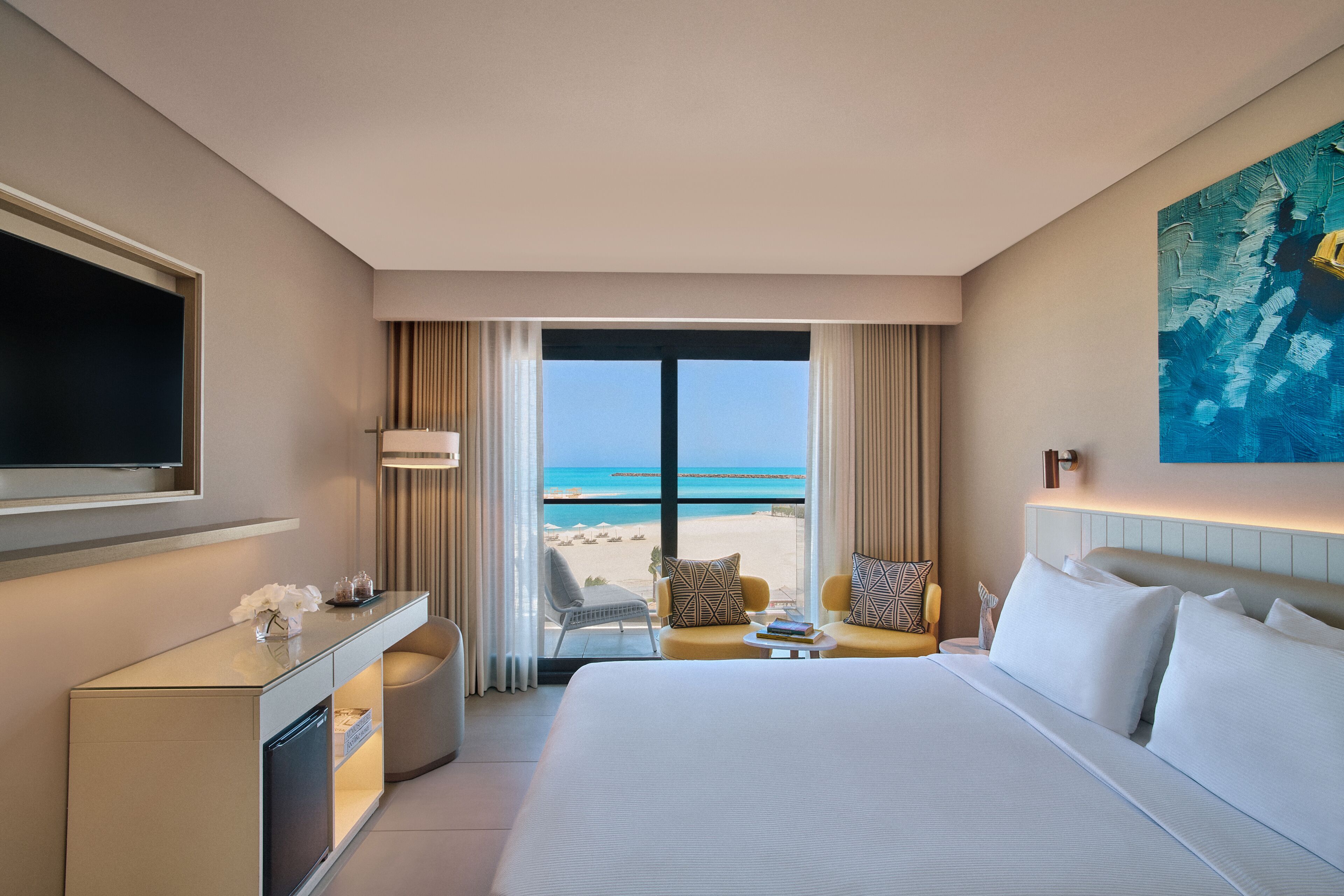 A hotel room with a bed, a desk, a TV, a view of the beach, and a painting on the wall.