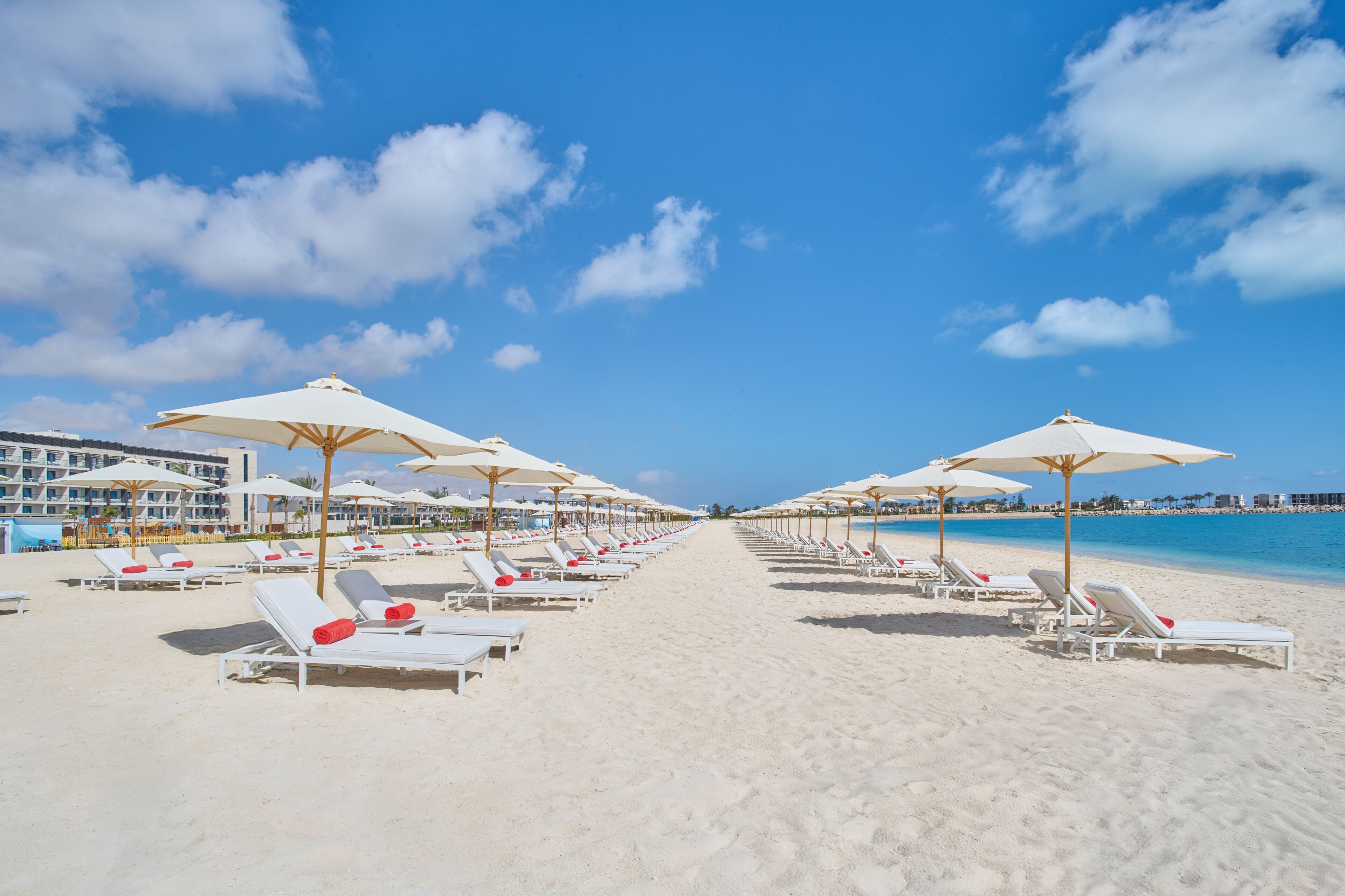 A beach with rows of sun loungers and umbrellas, a clear sky, and a hotel in the background.