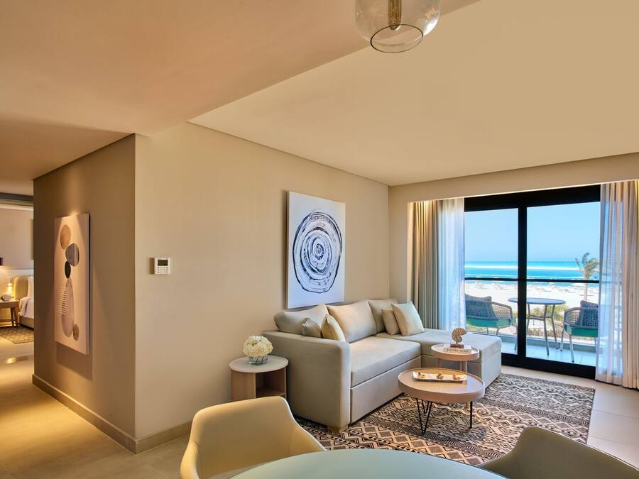 A modern hotel room with a balcony overlooking the beach, a sofa, a coffee table, and a painting on the wall.