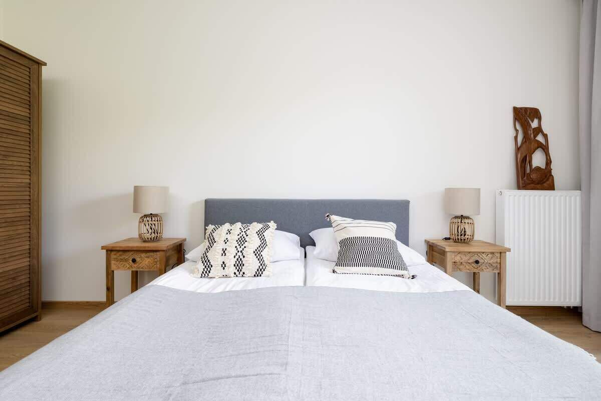 Ein ordentlich bezogenes Bett mit zwei Kissen, ein Nachttisch mit einer Lampe und ein Heizkörper an der Wand.
