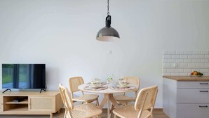Zona pranzo con tavolo rotondo, quattro sedie in vimini, una televisione su un mobile in legno e angolo cottura con parete rivestita in piastrelle bianche e una ciotola di frutta.