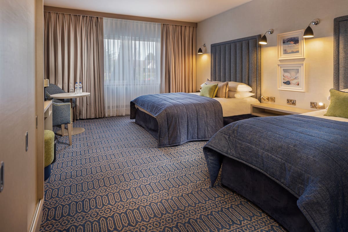 Habitación de hotel con dos camas, un escritorio, una silla, una mesita y un ventanal con cortinas.