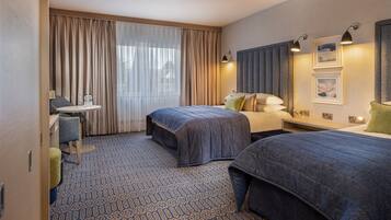 A hotel room with two beds, a desk, a chair, a small table, and a window with curtains.