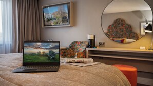 Habitación de hotel con una computadora portátil sobre la cama, un cuadro en la pared, un espejo redondo y un cojín estampado con motivos florales.