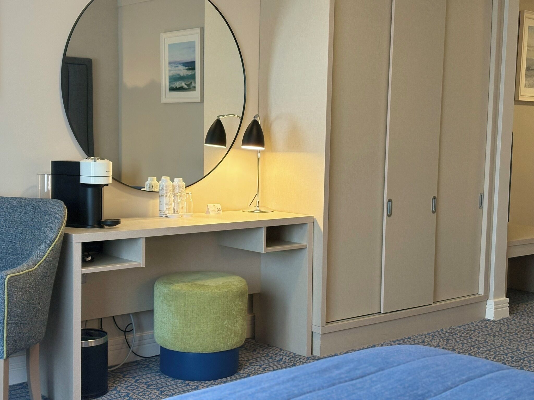 A hotel room with a bed, a desk with a mirror, a lamp, a chair, a wardrobe, and a small stool.