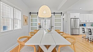 Une cuisine moderne avec des meubles blancs, un réfrigérateur en acier inoxydable et un coin repas comprenant une table blanche et des chaises en osier.