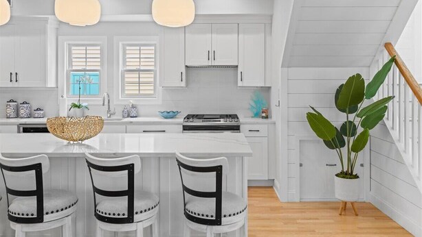 Une cuisine moderne avec des meubles blancs, un îlot central doté de tabourets de bar, des luminaires suspendus et une plante en pot.
