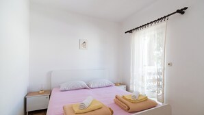 A neatly made bed with pink bedding and two folded towels, a bedside table with a lamp, and a window with sheer curtains. - Apartments Pinija - (Supetar)