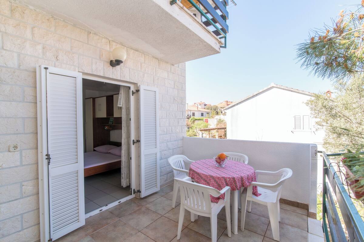 Un balcón con una mesa y sillas, una cama con contraventanas y vistas a edificios cercanos.