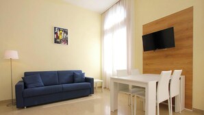 A modern living room with a blue sofa, a white dining table with chairs, a flat-screen TV mounted on a wooden wall, and a floor lamp. - Romantic Venetian Apartment with Grand Canal Views and High Ceilings (Venezia)