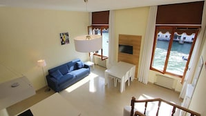A room with a blue sofa, a dining table with chairs, a TV, and a view of the sea through large windows. - Romantic Venetian Apartment with Grand Canal Views and High Ceilings (Venezia)