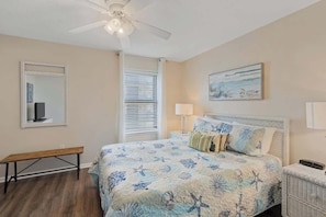 A bedroom with a bed, a bench, a ceiling fan, a window with blinds, and a wall-mounted TV.
