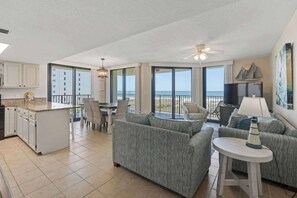 A spacious living area with a kitchen, dining table, and sofas, large windows offering a view of the ocean.