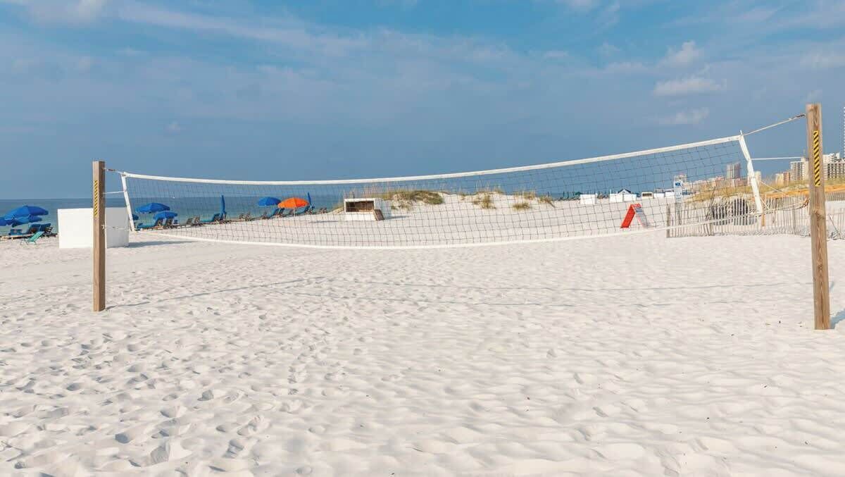 A beach volleyball net on a sandy beach with umbrellas and beach chairs in the background.