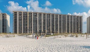 Un hotel frente a la playa con varios edificios altos, una playa de arena en primer plano y un cielo despejado.