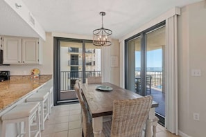A dining area with a wooden table and wicker chairs, a kitchen with white cabinets, and a balcony with a view.